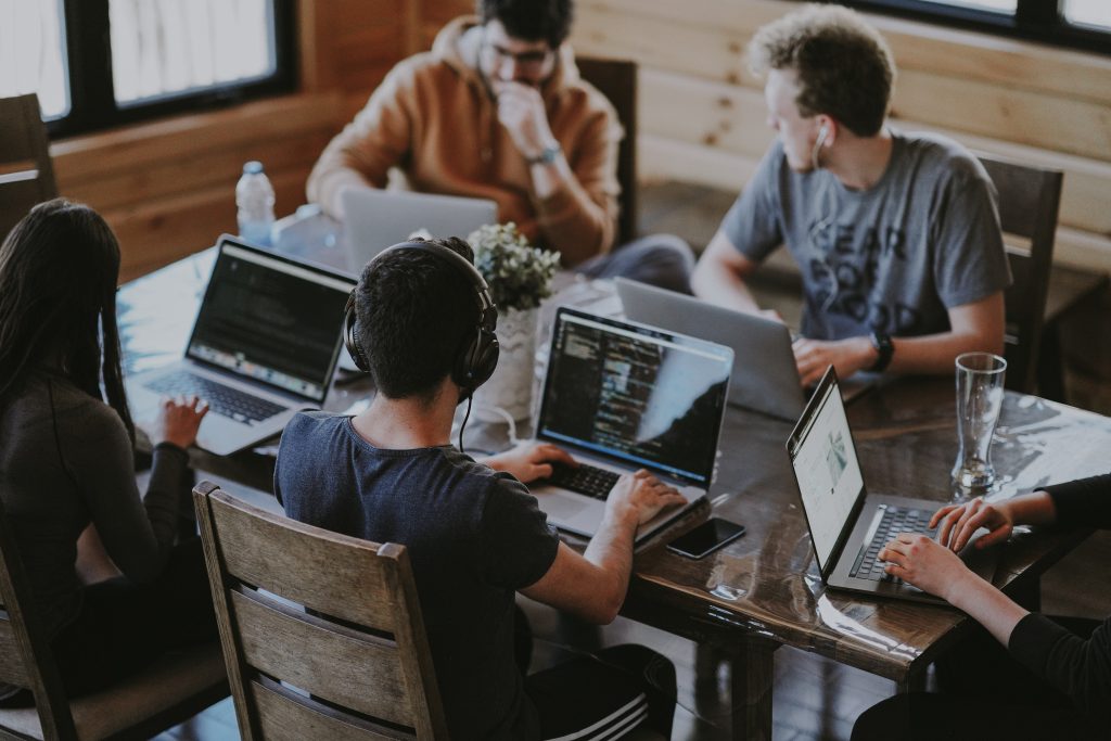 A group of young adults sits around a wooden table working on laptops in a collaborative workspace. Some are wearing headphones, and one screen shows lines of code, indicating a tech-focused environment. The image reflects the role of digital technology in modern team settings, highlighting the impact of technology in K12-Education and beyond.