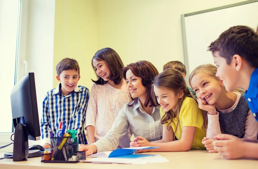 A female teacher sits at a desk surrounded by a group of smiling elementary school children, all looking at a computer monitor together. The scene represents the integration of digital technology in K12-education, highlighting the use of technology as a collaborative learning tool in a classroom setting.