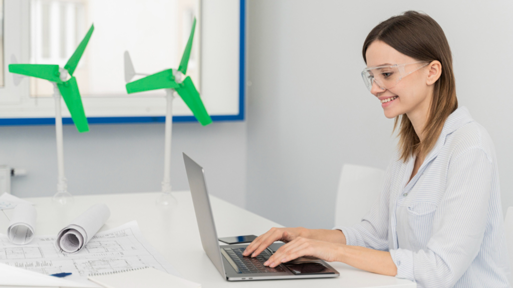 A smiling woman wearing safety glasses types on a laptop at a white desk with wind turbine models and blueprints. The image reflects how professionals in engineering and energy sectors benefit from a Fiber Internet Connection for real-time analysis and system management.