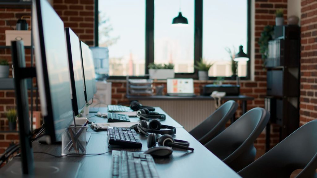 A modern office with multiple computer workstations, headsets, and ergonomic chairs. The space is well-lit with large windows and a brick wall background, representing a tech-savvy workspace ideal for Hosted PBX Solutions and Business phone solutions.