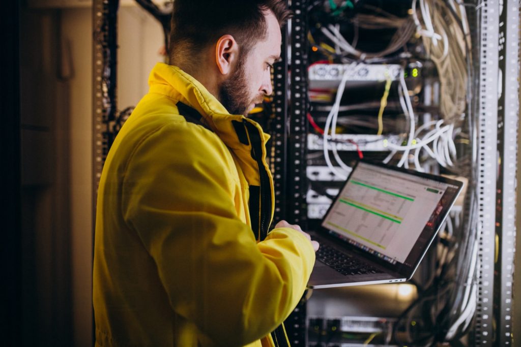 A network technician wearing a bright yellow jacket works on a laptop while standing in front of a server rack filled with cables and equipment. The scene highlights the importance of securing IT infrastructure against ransomware attacks.