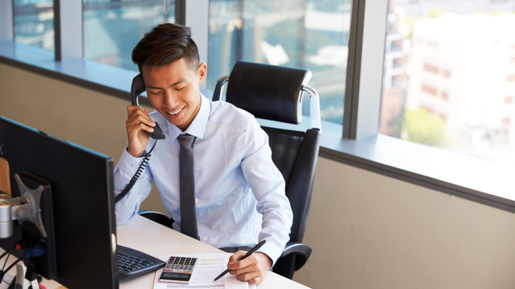 Young professional in a dress shirt and tie smiles while speaking on a corded office phone at a desk in a modern, sunlit office. He is simultaneously writing on a document beside a calculator and computer monitor. This image illustrates the effectiveness and multitasking capabilities supported by Enterprise VoIP solutions in a corporate environment.