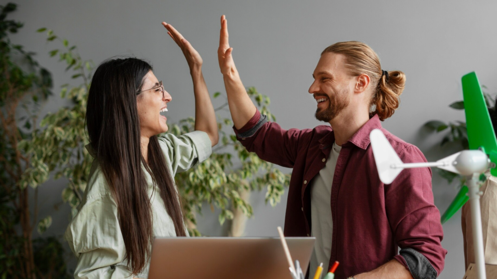 A smiling man and woman give each other a high five in a bright, plant-filled office space, celebrating a successful moment in front of a laptop. A small wind turbine model is visible nearby, suggesting innovation and sustainability. The scene symbolizes teamwork and success in promoting cybersecurity awareness within a positive work environment.