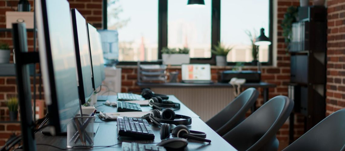 A modern office with multiple computer workstations, headsets, and ergonomic chairs. The space is well-lit with large windows and a brick wall background, representing a tech-savvy workspace ideal for Hosted PBX Solutions and Business phone solutions.