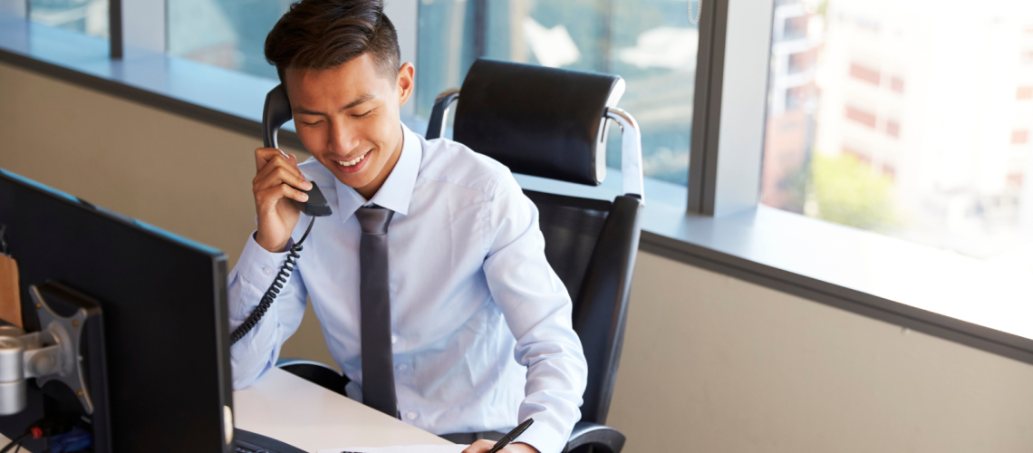 Young professional in a dress shirt and tie smiles while speaking on a corded office phone at a desk in a modern, sunlit office. He is simultaneously writing on a document beside a calculator and computer monitor. This image illustrates the effectiveness and multitasking capabilities supported by Enterprise VoIP solutions in a corporate environment.
