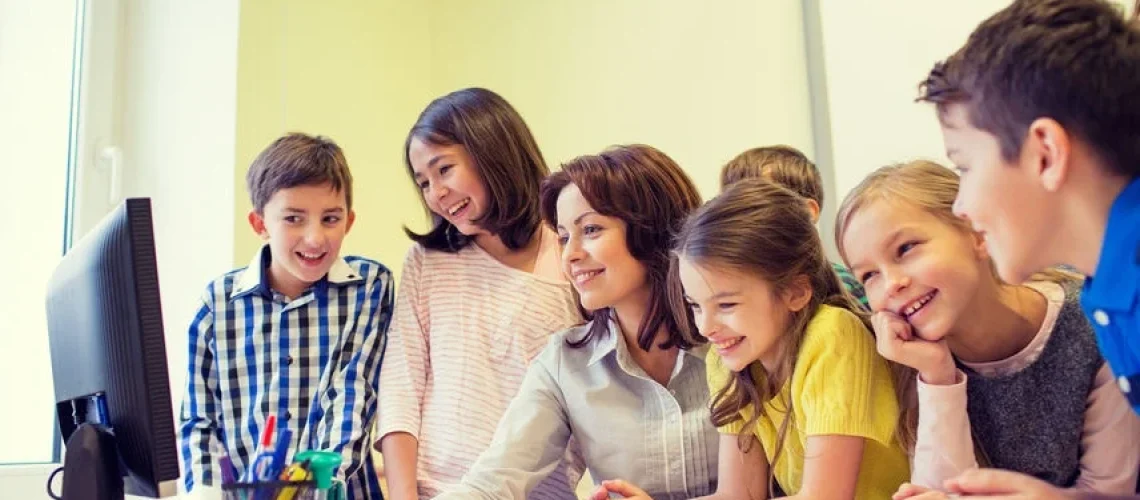 A female teacher sits at a desk surrounded by a group of smiling elementary school children, all looking at a computer monitor together. The scene represents the integration of digital technology in K12-education, highlighting the use of technology as a collaborative learning tool in a classroom setting.