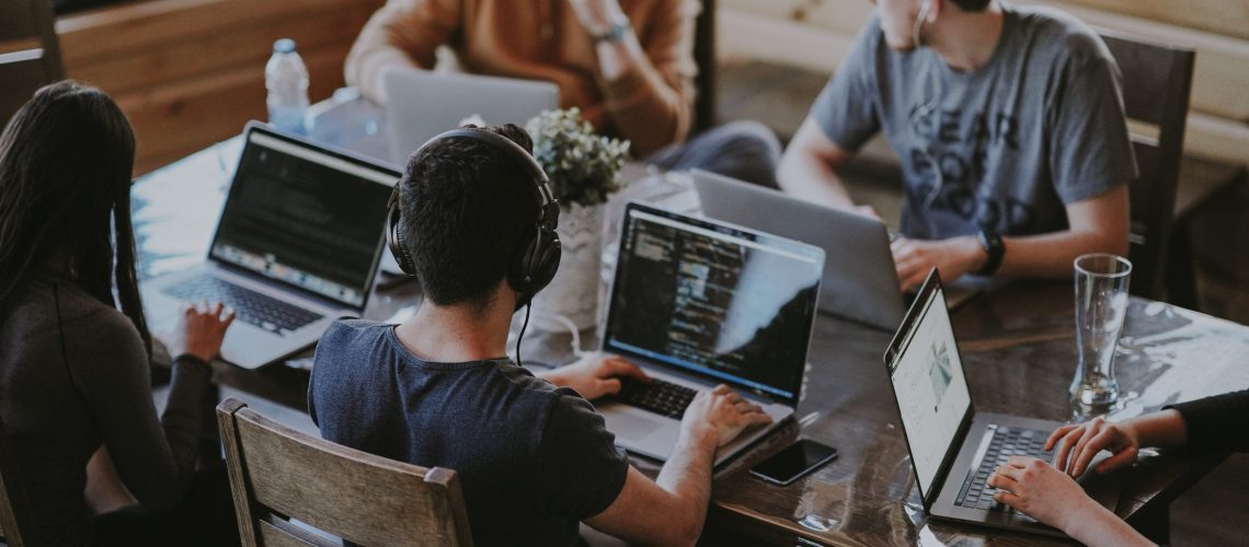 A group of young adults sits around a wooden table working on laptops in a collaborative workspace. Some are wearing headphones, and one screen shows lines of code, indicating a tech-focused environment. The image reflects the role of digital technology in modern team settings, highlighting the impact of technology in K12-education and beyond.